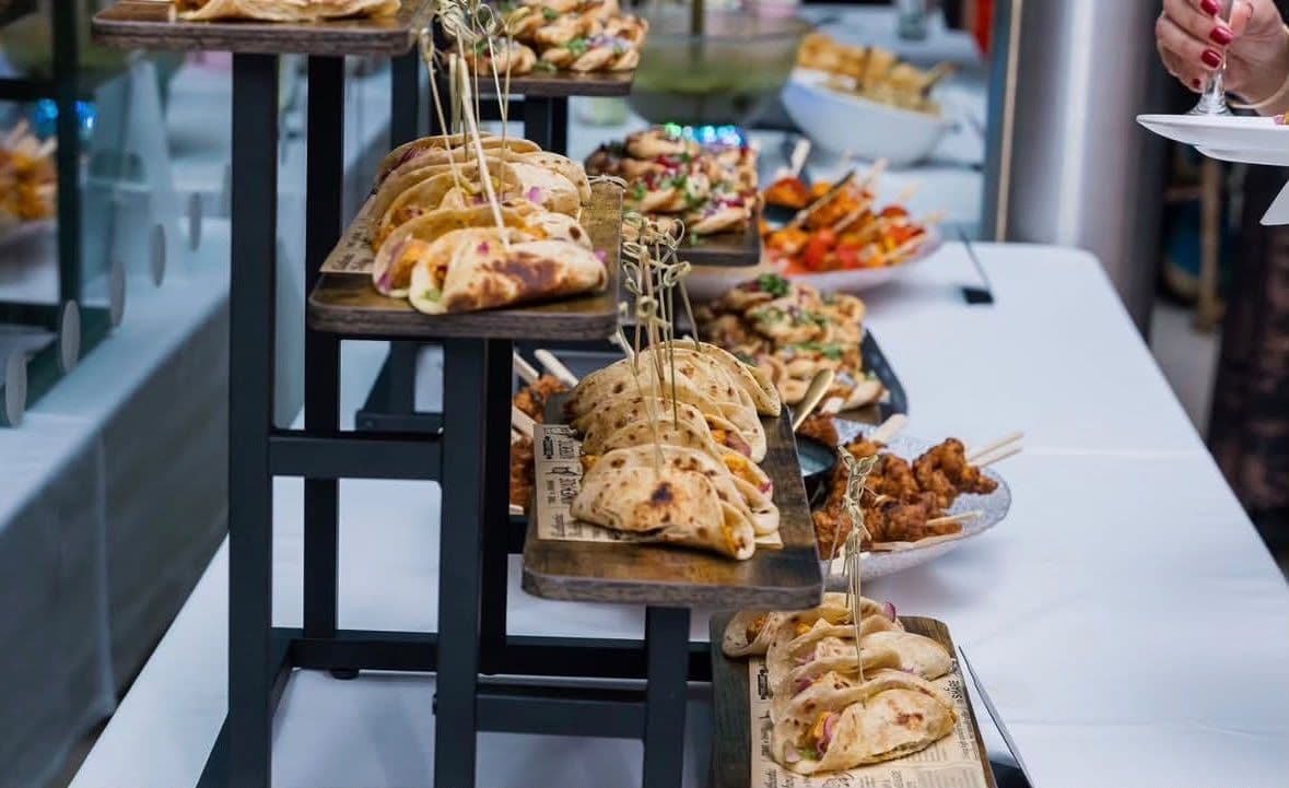 Rows of folded flatbread wraps with spiced fillings and red onion on a tiered wooden stand, with skewered bites and more dishes laid out along a white table behind